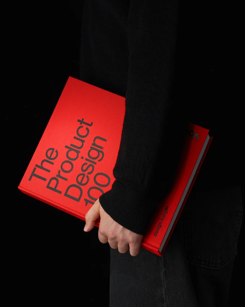 Uma pessoa de roupa preta carrega debaixo do braço o livro The Product Design 100, de capa vermelha com o título em letras pretas e a assinatura design burger na lombada