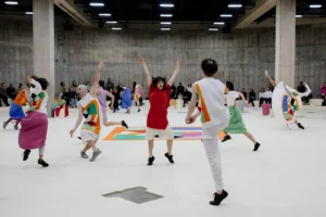 Bailarinos da Dresden Frankfurt Dance Company em roupas plissadas coloridas dançam no tanque sul da Tate Modern, com a recriação de The Snail em roupas no chão ao fundo e o público sentado junto às paredes de concreto