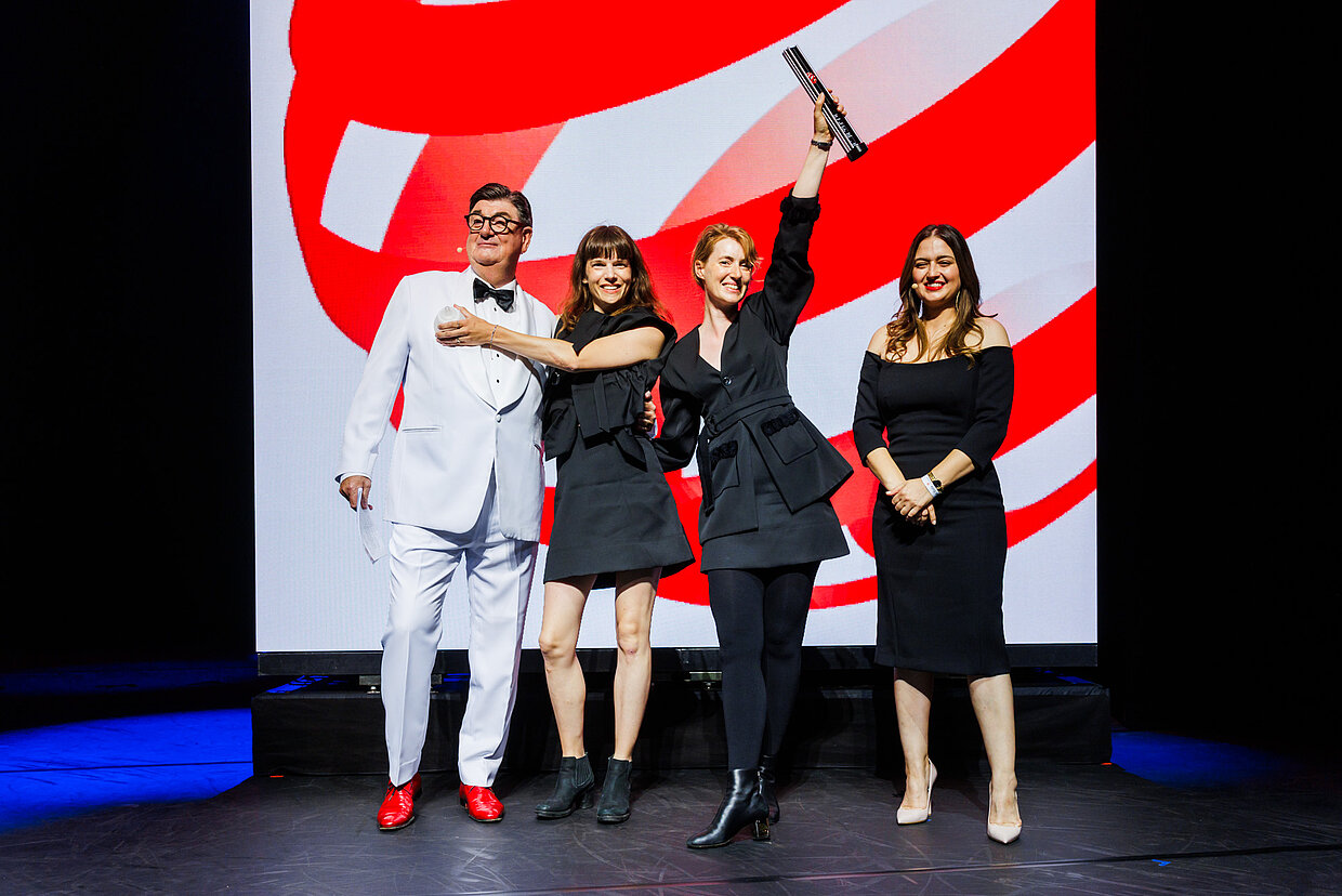 Quatro pessoas no palco da gala do Red Dot, uma delas erguendo o troféu, diante do símbolo vermelho e branco do prêmio