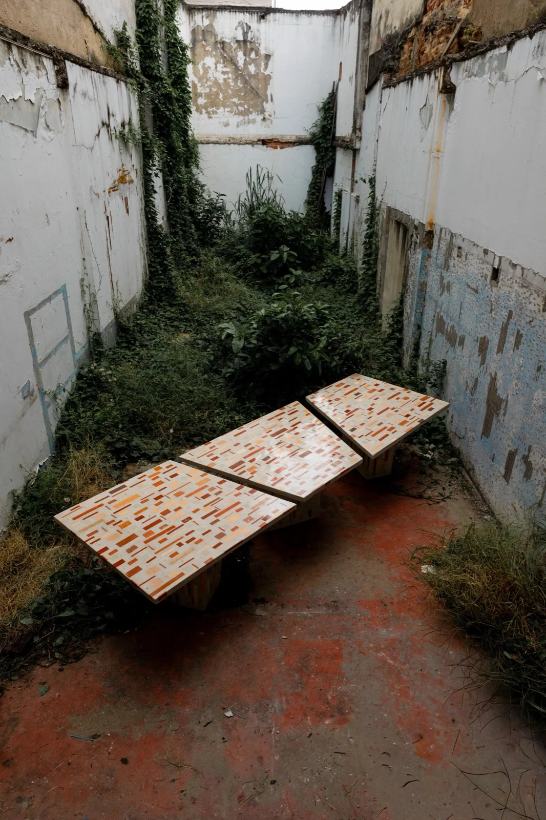Três módulos da mesa Dominó, com tampos de cerâmica em tons de laranja e terracota, num terreno murado em ruína tomado por mato