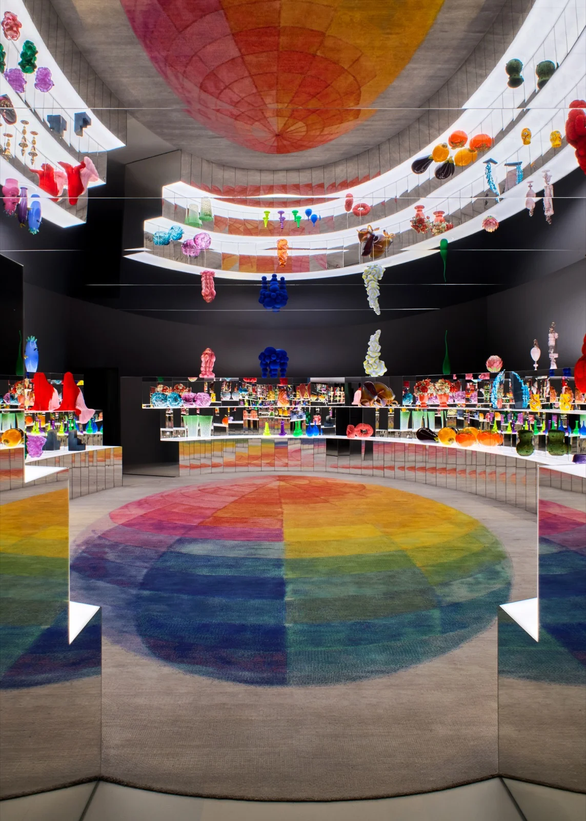 Sala com prateleiras espelhadas cheias de objetos coloridos de vidro e resina, teto espelhado e tapete com círculo de cores do arco-íris