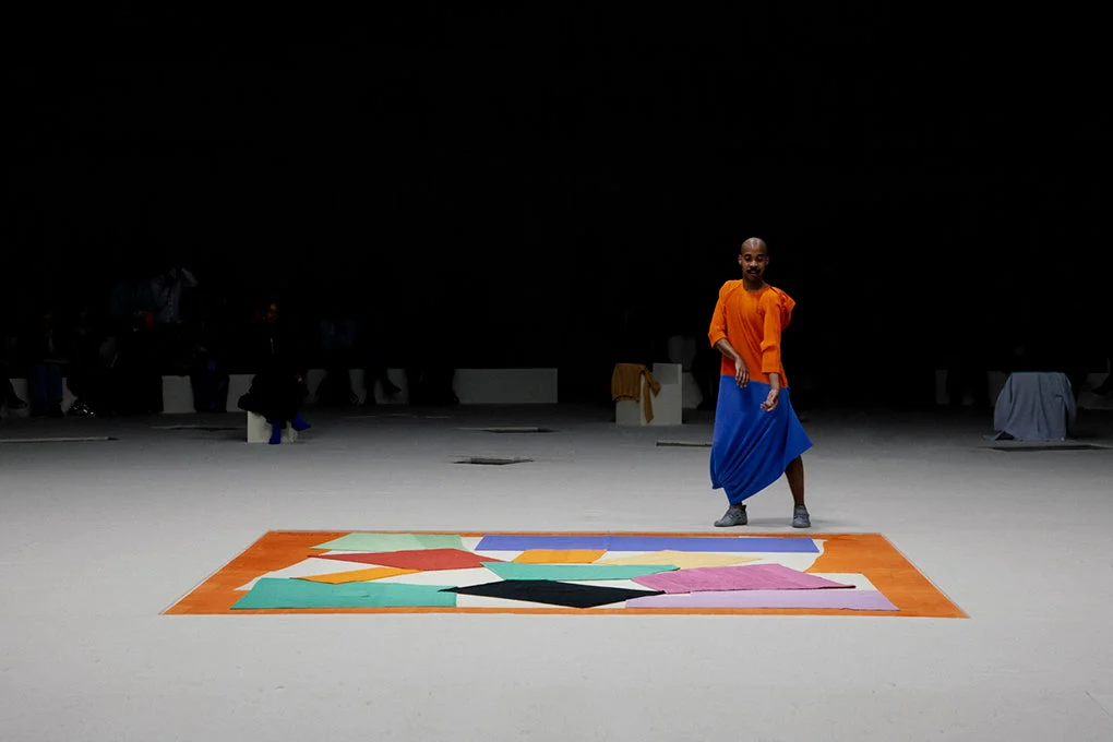 Recriação de The Snail feita de roupas plissadas estendidas no chão de feltro branco, com borda laranja, ao lado de um performer de blusa laranja e saia azul no tanque escuro