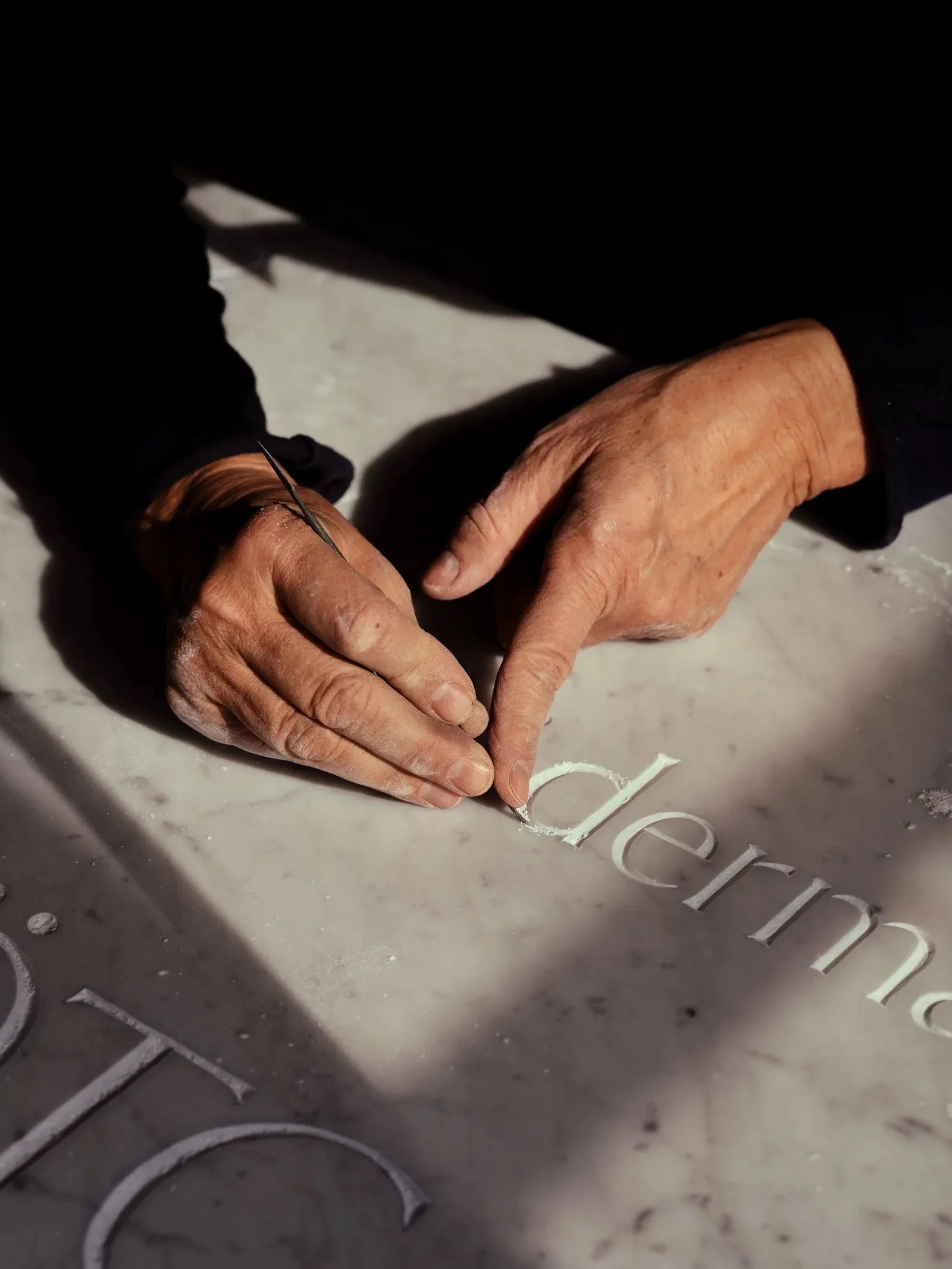 Duas mãos seguram um cinzel fino sobre uma placa de mármore, terminando a talha de letras minúsculas com serifa