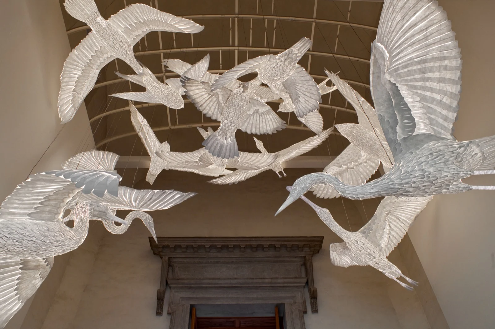 Garças e outras aves de papel branco iluminadas, suspensas sob a abóbada da escadaria do refeitório palladiano
