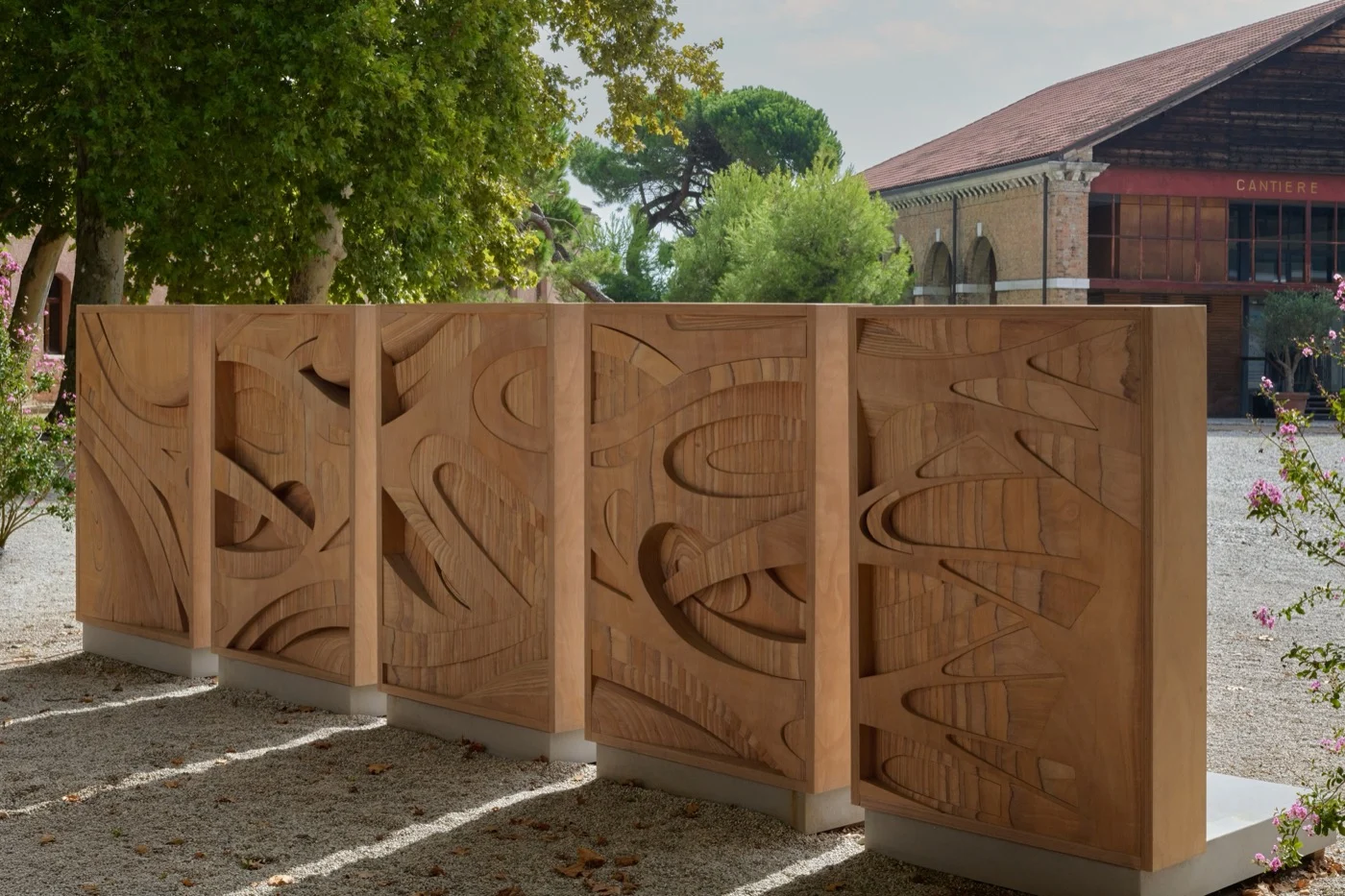 Painéis de madeira curvada com relevos orgânicos enfileirados num pátio de cascalho, ao lado de árvores e de um galpão escrito Cantiere